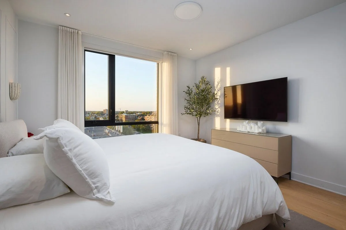 A modern bedroom with a large bed, white bedding, a window with white curtains, a TV mounted on the wall, a dresser, and a potted plant next to the window.