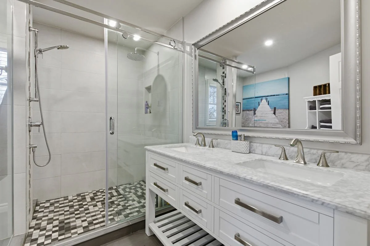 Modern bathroom with a glass shower enclosure, white vanity with marble countertop, large mirror, and nautical artwork