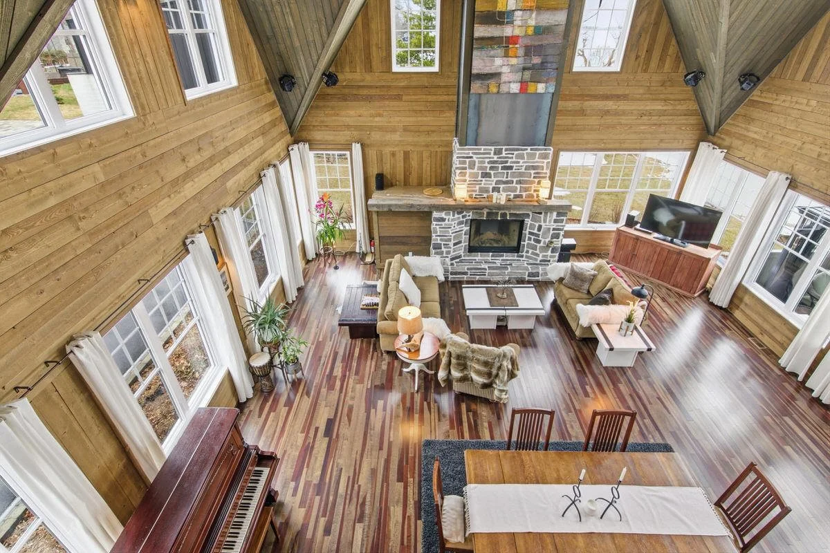 A spacious living room with wooden walls and ceiling, large windows with white curtains, a stone fireplace, and hardwood floors. The room has sofas, a coffee table, a TV on a wooden stand, plants, and decorative items.