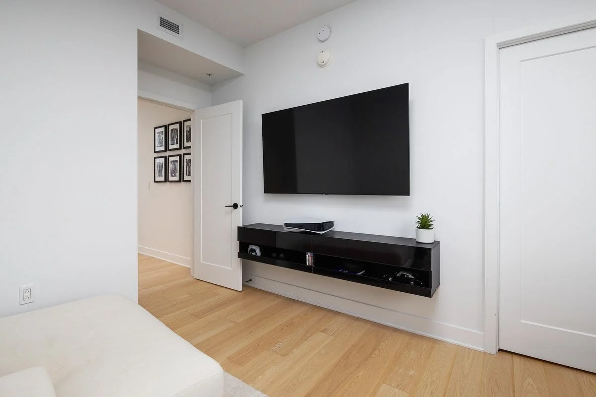 Minimalist living room with a mounted flat-screen TV, black entertainment console, small potted plant, and framed black-and-white photos on the wall.