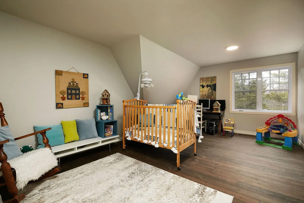 A child's nursery with a crib, a play area, toys, and a small computer desk under an angled ceiling with a window.