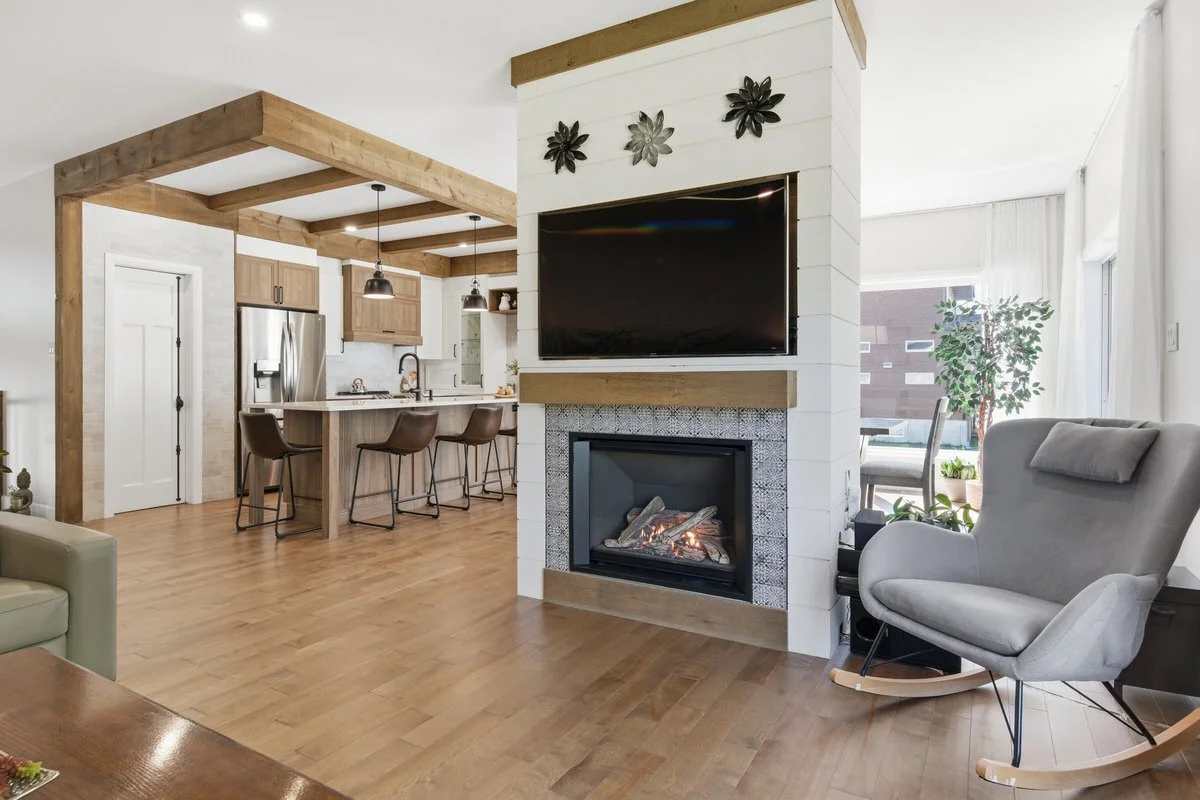 Living room with a fireplace, a TV mounted above it, a gray rocking chair, and large windows with plants outside. The room features a modern open kitchen with a breakfast bar and pendant lighting.