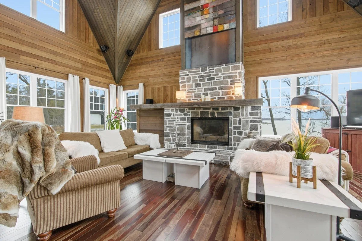 A cozy living room with wood-paneled walls and ceiling, large windows, a stone fireplace, and various comfortable seating including a striped armchair, a beige couch, and a white bench. Decor includes a fur throw, potted plants, candles, and a colorf