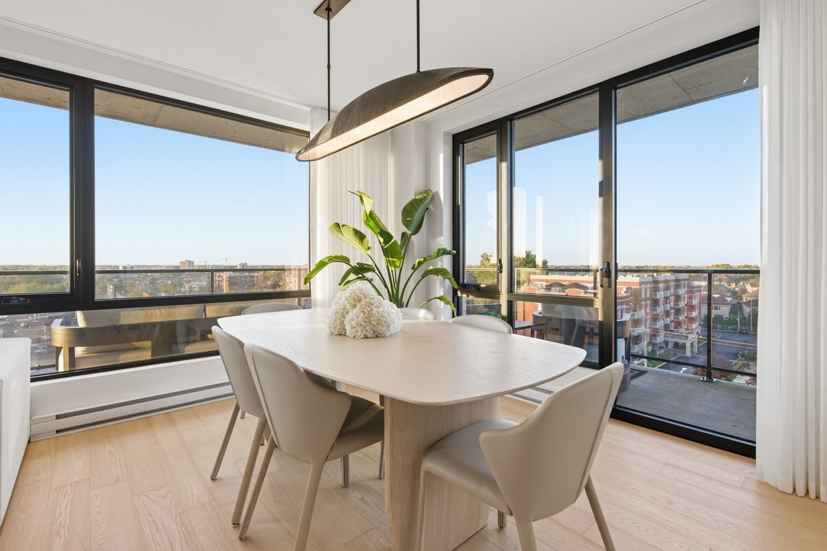 Modern dining room with a white table, four beige chairs, a potted plant, and large glass sliding doors leading to a balcony with a city view.