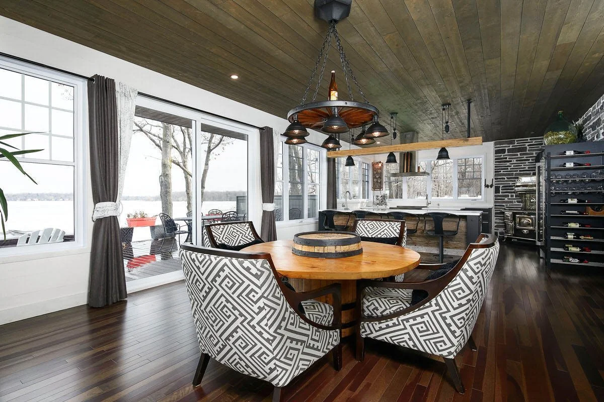Open-concept dining area with round wooden table, six patterned chairs, sliding glass doors leading to an outdoor deck, and a kitchen with bar stools and windows in the background.