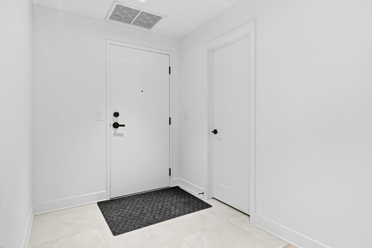 A minimalist white entryway with a front door, a vent on the ceiling, and a smaller door on the right, all with black handles, and a dark gray doormat on light-colored tile flooring.