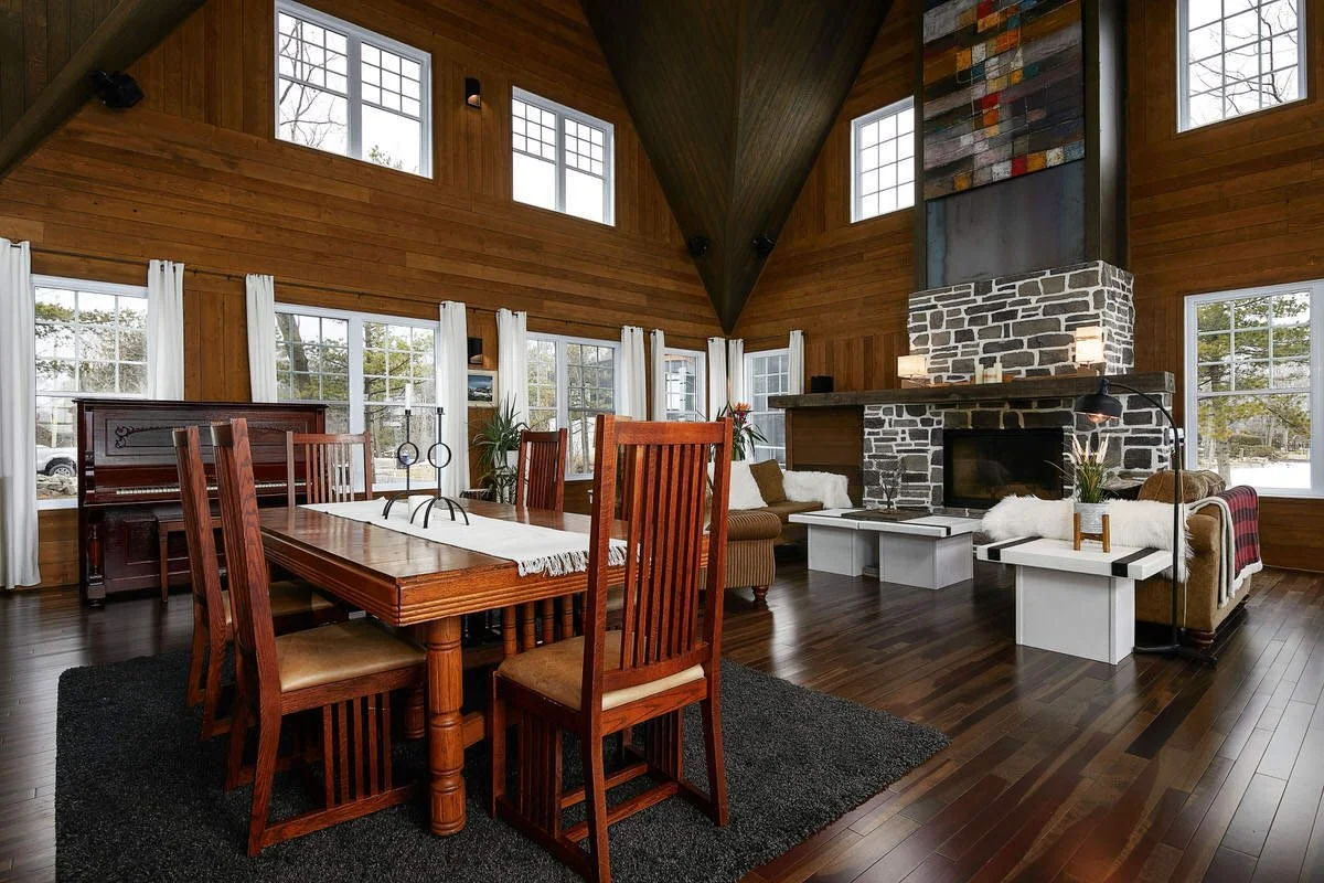 A spacious living and dining area with high vaulted wood-paneled ceilings, large windows with white curtains, and a hardwood floor. The dining table is made of wood with six chairs, and there is a black rug underneath. The living area features a ston