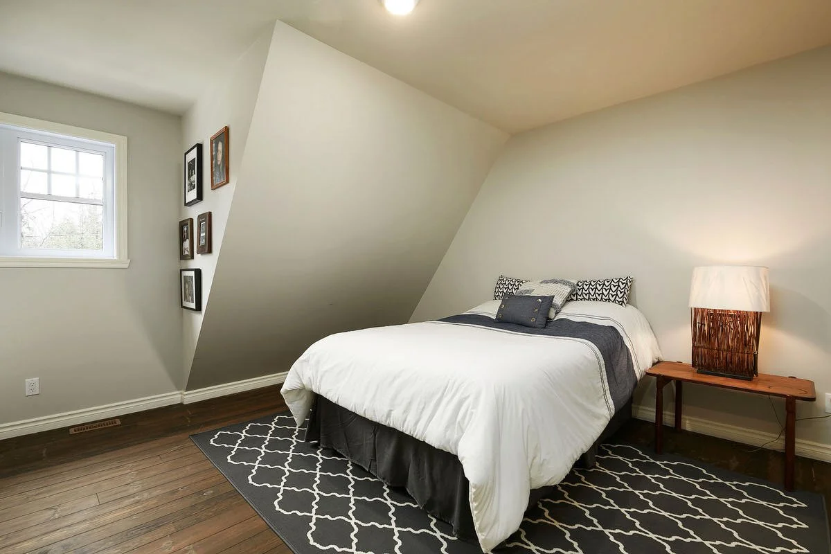 A bedroom with a bed, a lamp on a small wooden table, a black and white area rug, a window, and framed art on the walls.