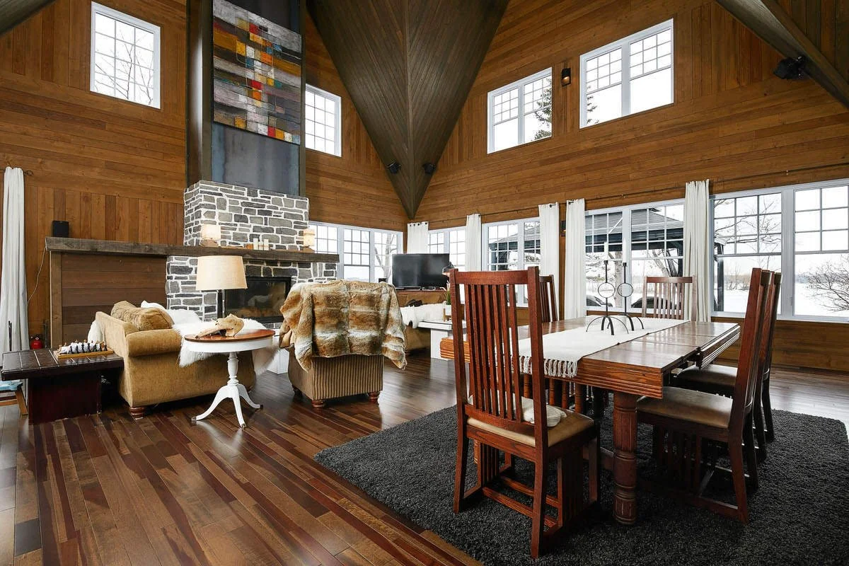 A spacious living and dining room with wood-paneled walls and ceiling, large windows with white curtains, a stone fireplace, and wooden dining table with chairs.