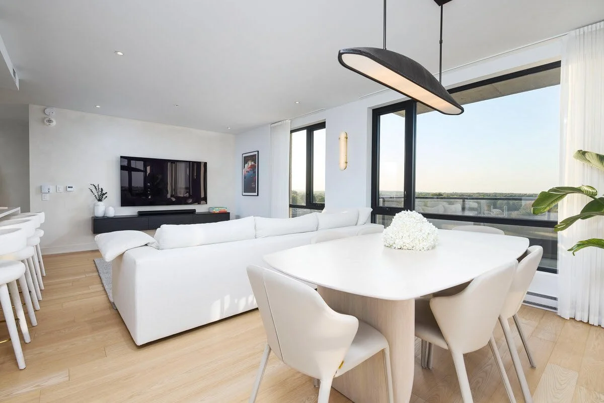 Modern living room with large windows, white sofa, wall-mounted TV, and dining area with white chairs and a floral centerpiece.