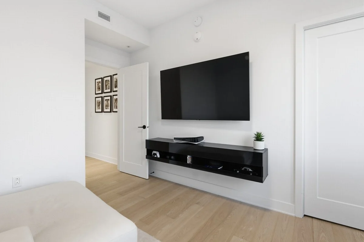 Modern living room with a mounted flat-screen TV above a black floating media console, a small potted plant, and framed artwork on the hallway wall.