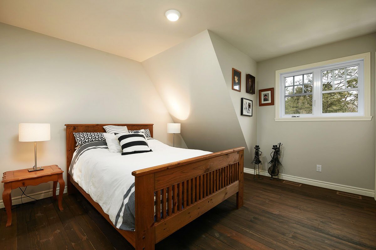 Bedroom with a wooden bed, white bedding, black and white pillows, wall-mounted lights, framed pictures on the wall, and a window showing trees outside.