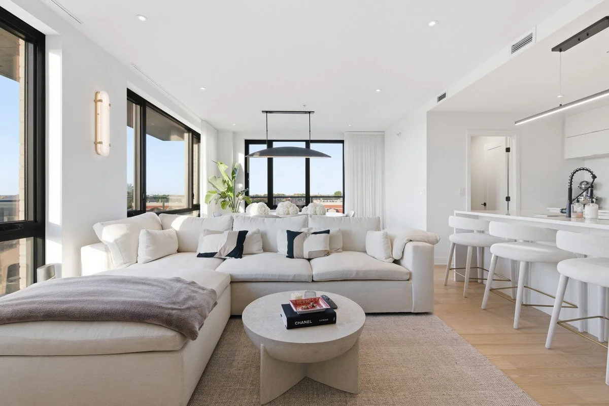 Modern white living room with large sectional sofa, coffee table, bar stools, mountain view through windows, and minimal decor.