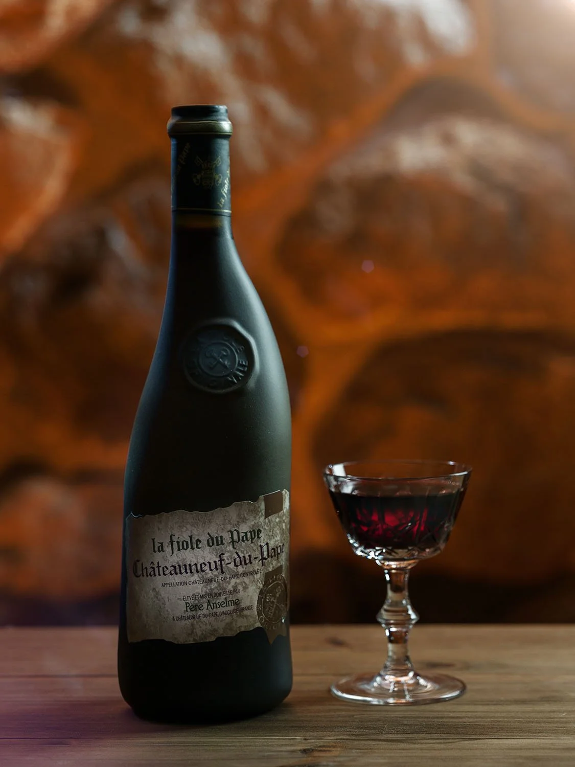 A bottle of La Fiole du Pape wine on a wood table against a stone wall.