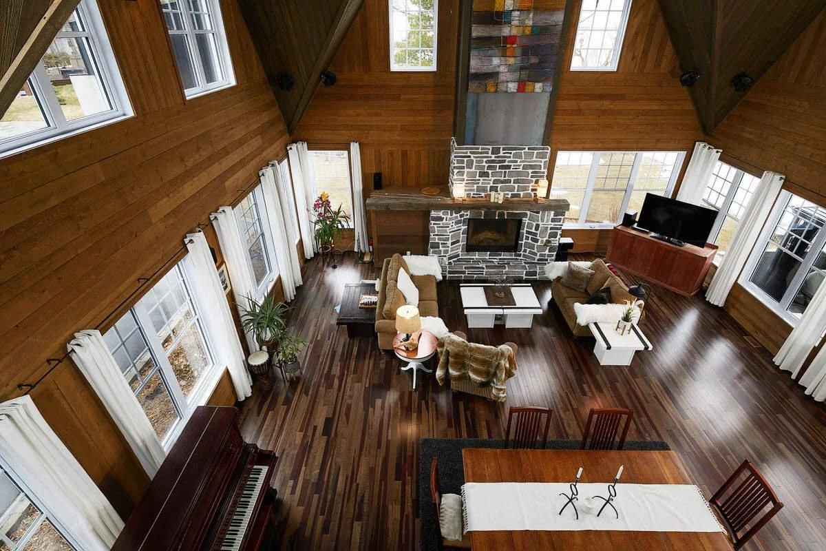 A spacious living room with wooden walls and ceiling, multiple large windows with white curtains, and dark wooden floors. There is a stone fireplace, a flat-screen TV on a wooden cabinet, a brown sofa, an armchair with a blanket, a coffee table, and 