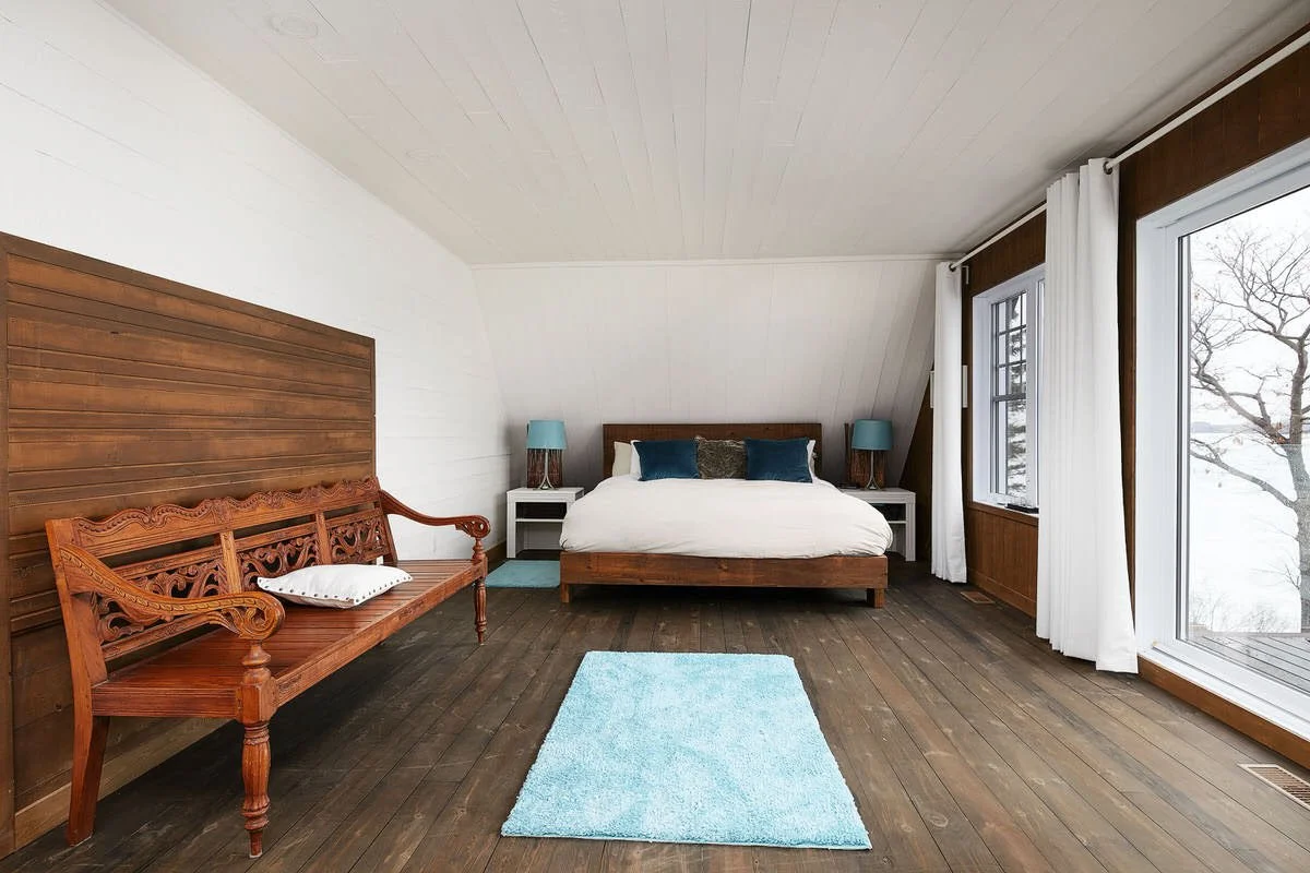 A bedroom with wooden floors, a large bed with white bedding and blue pillows, two white nightstands with blue lamps, an ornate wooden bench with a pillow, and large windows with white drapes showing snow outside.