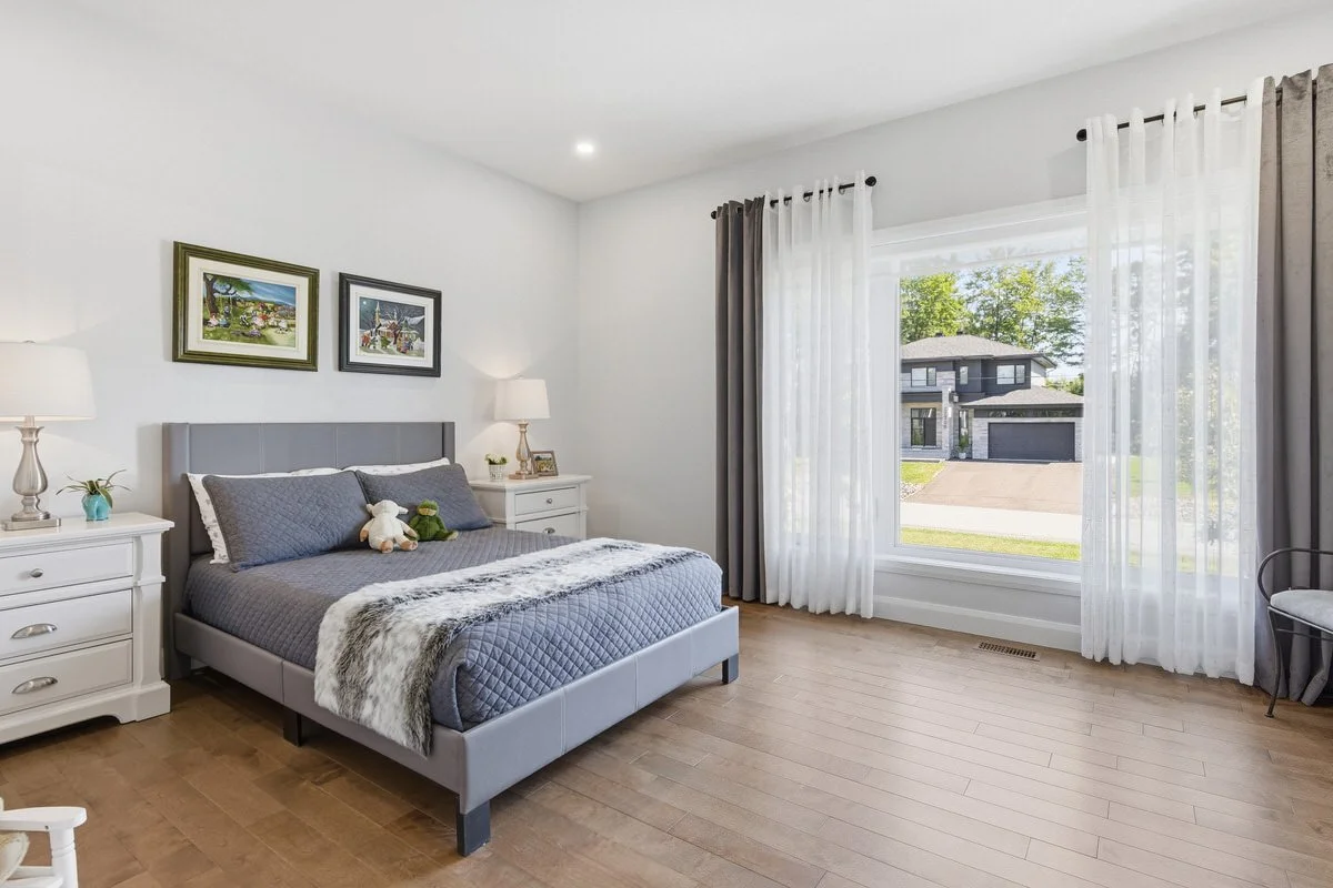 A bedroom with a large gray bed, nightstands with lamps, family photos, and stuffed animals, light wood flooring, and large windows with white and dark curtains showing a view of a suburban street.