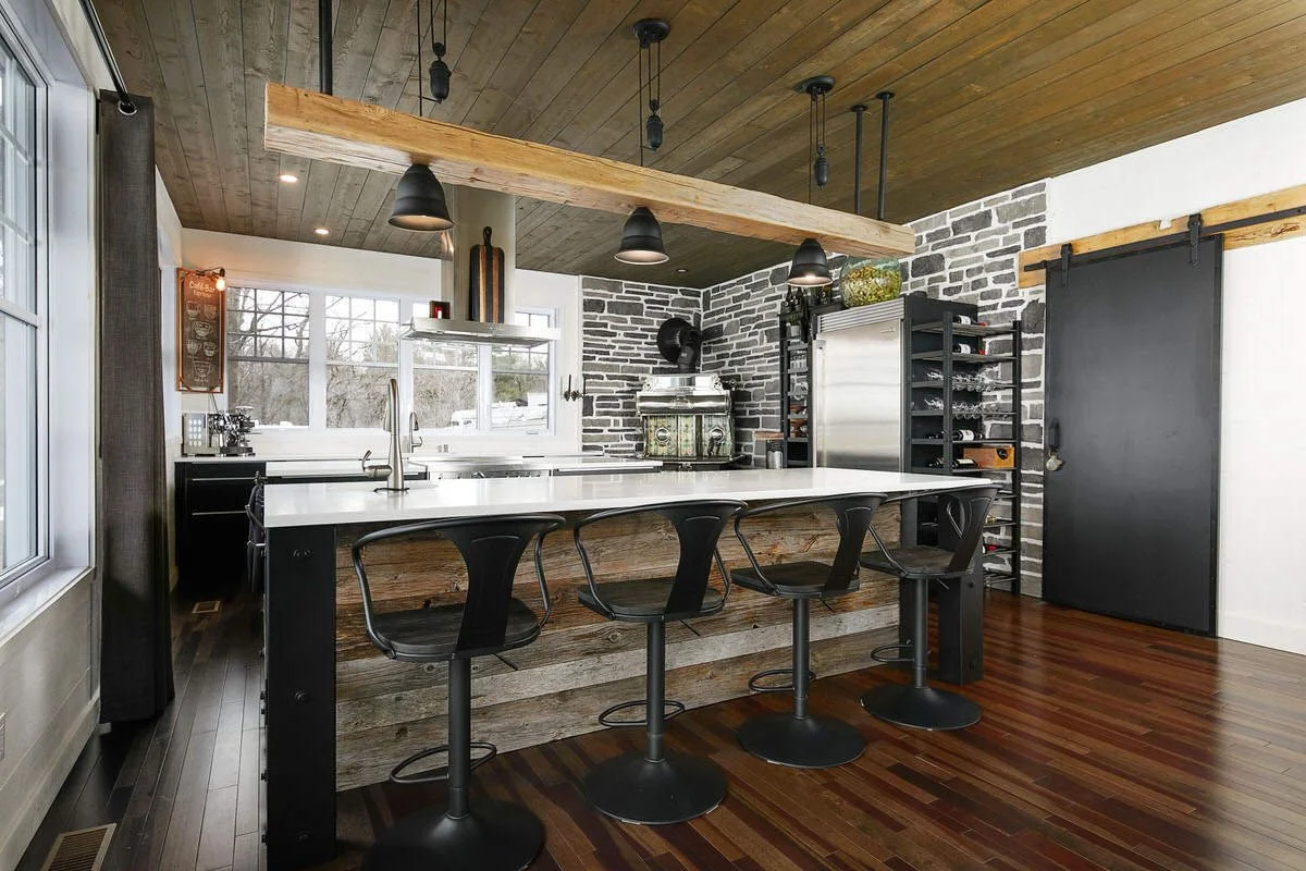 Modern kitchen with a farmhouse style island, black bar stools, white countertops, brick walls, and a wooden ceiling.