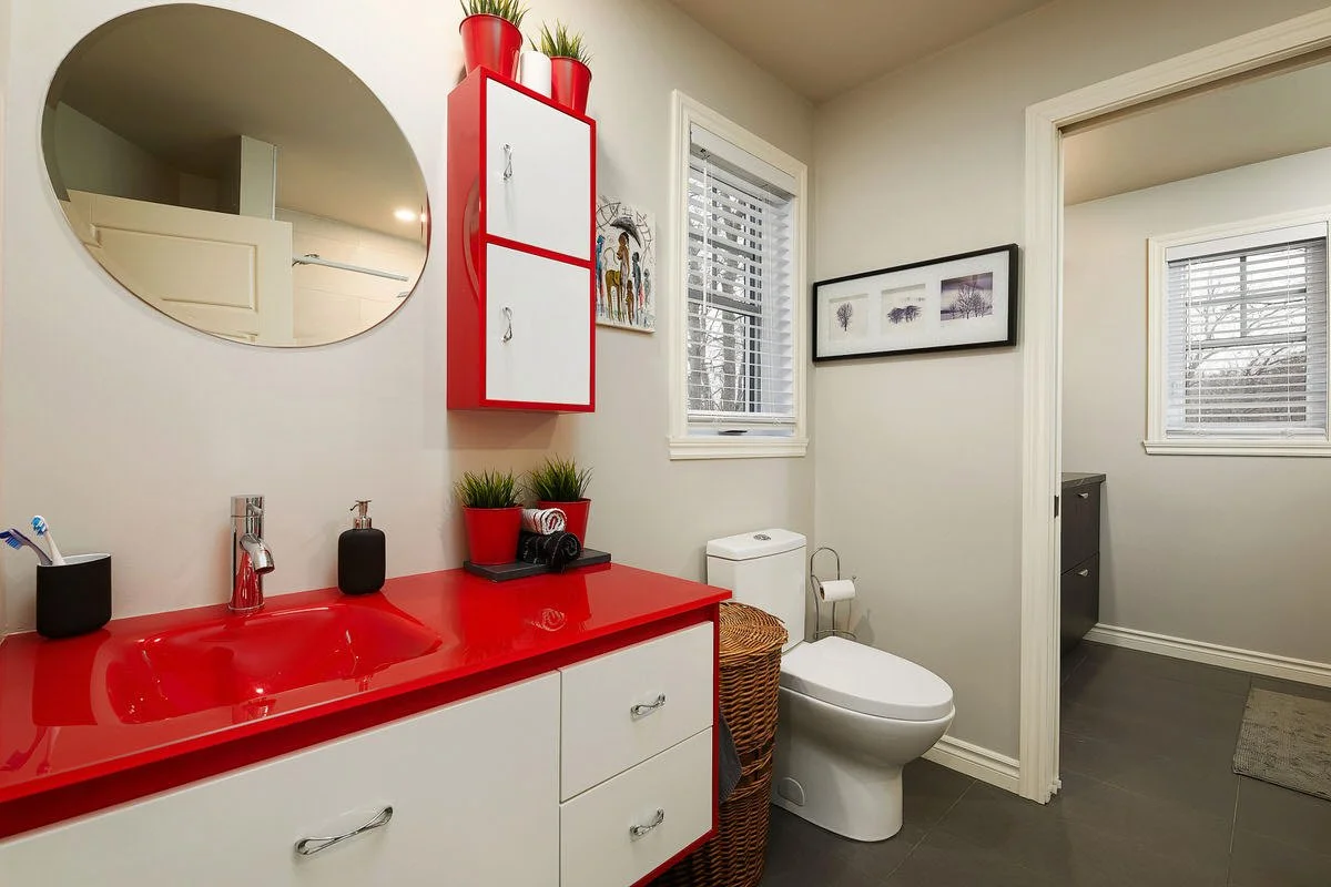 Bathroom with a red countertop sink, black toothbrush holders, potted plants, a wicker laundry basket, a framed black-and-white artwork, and a window with blinds.