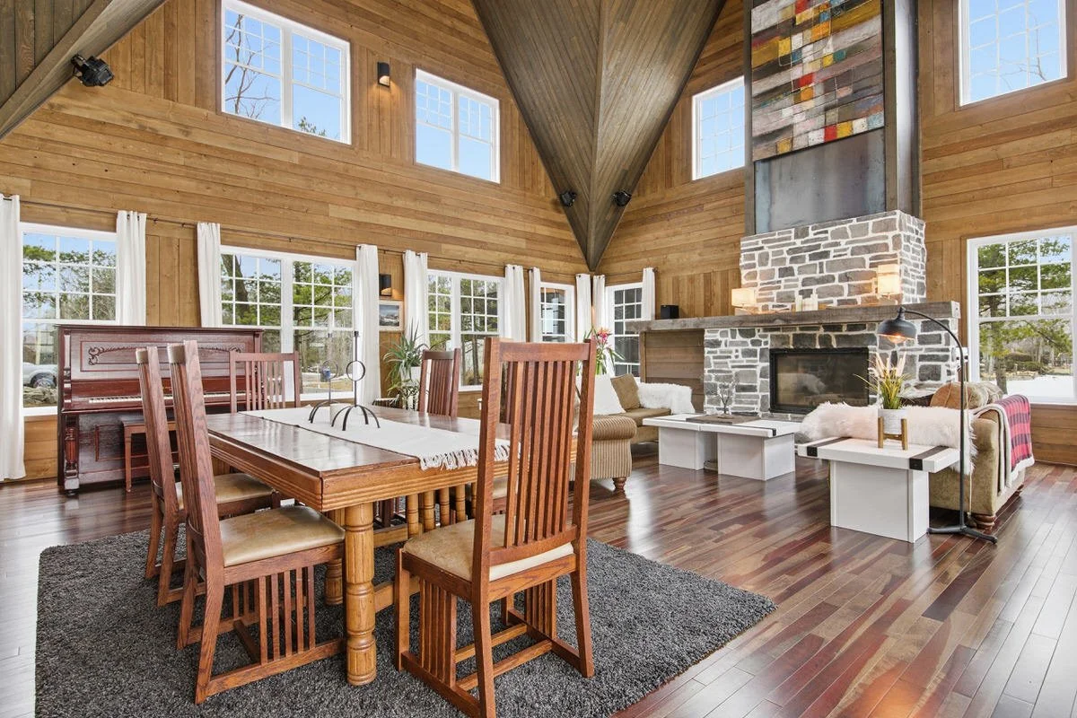 A spacious living room with a high, vaulted wooden ceiling, large windows with white curtains, a stone fireplace, and wooden flooring. The room has a dining table with six wooden chairs, a cozy seating area with sofas and armchairs, and various decor