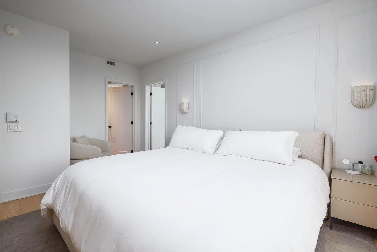 A clean, minimalistic bedroom with a large white bed and matching pillows, a beige nightstand with a lamp and a small plant, and white walls with wall sconces. There is a doorway and a small sitting area visible in the background.