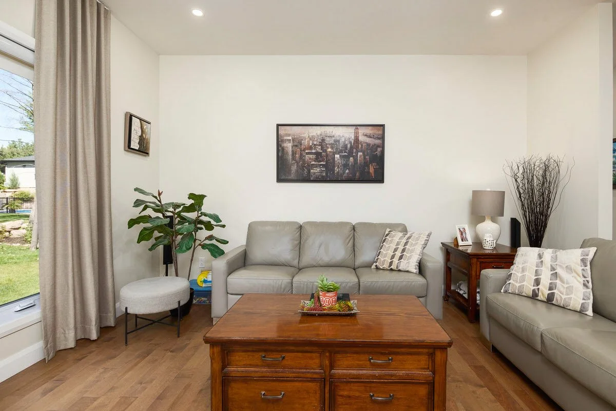 Living room with two beige leather sofas, a wooden coffee table with a small plant, a side table with a lamp, a framed cityscape picture on the wall, and a large window with beige curtains showing an outdoor yard.