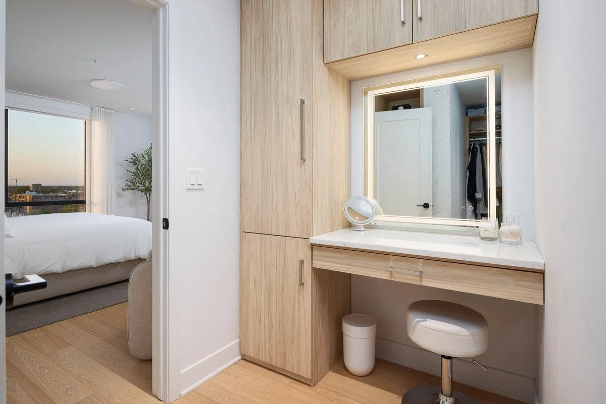 View of an interior bedroom and a small vanity area with a mirror, stool, and a white lamp. The bedroom has a bed, window, and cityscape view outside.