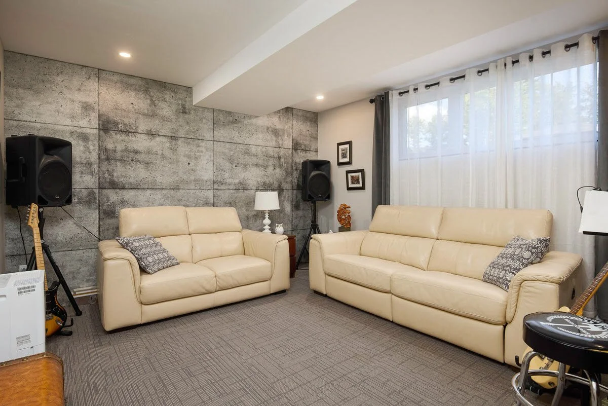 Living room with two cream-colored leather sofas, gray patterned pillows, a gray carpet, a window with sheer white curtains and gray drapes, a concrete accent wall, and musical equipment including speakers, guitars, and a microphone stand.