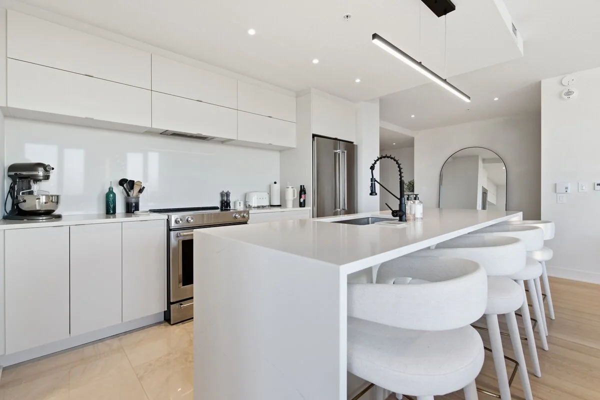 Modern white kitchen with a large island, bar stools, stainless steel appliances, and minimal decor.