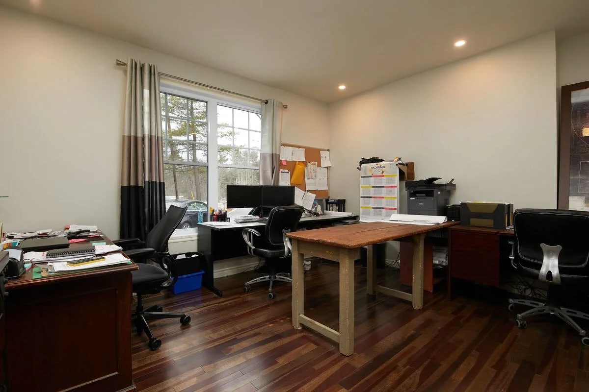 Office room with multiple desks, chairs, and office supplies, window with curtains, and hardwood floor.