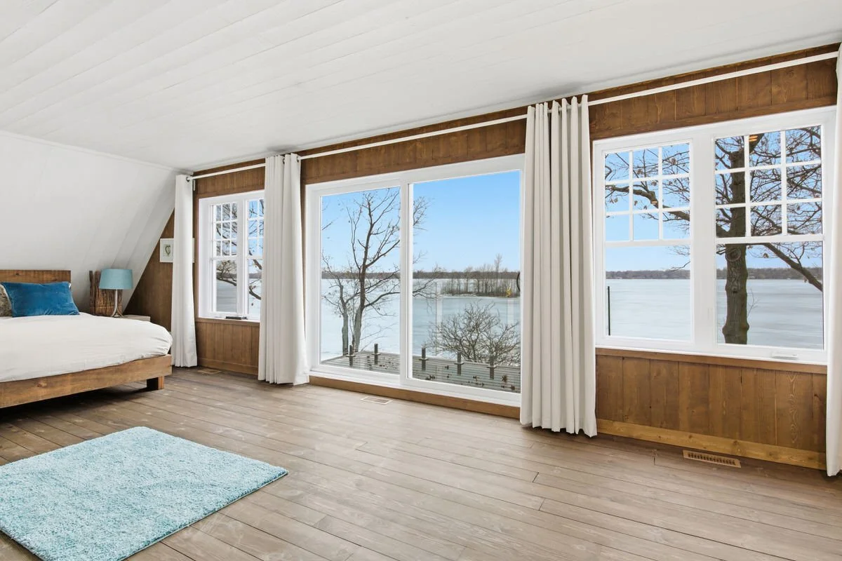 Bright bedroom with large windows overlooking a lake, wooden walls, white curtains, a bed with blue pillows, a small table with a lamp, and a light blue rug.