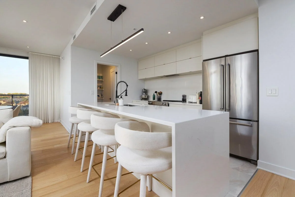 Modern kitchen with white cabinets, stainless steel refrigerator, and white island with barstools, adjacent to living room with large window and light-colored curtains.