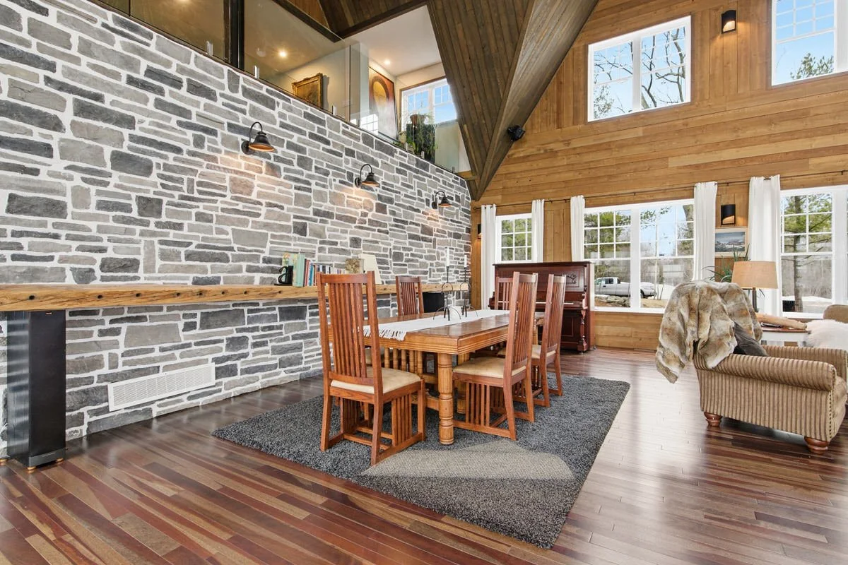 Interior of a spacious dining room with a modern rustic design, featuring a stone accent wall, wooden floors, a wooden dining table with six matching chairs, large windows with white curtains, and a cozy seating area with an armchair and a blanket.