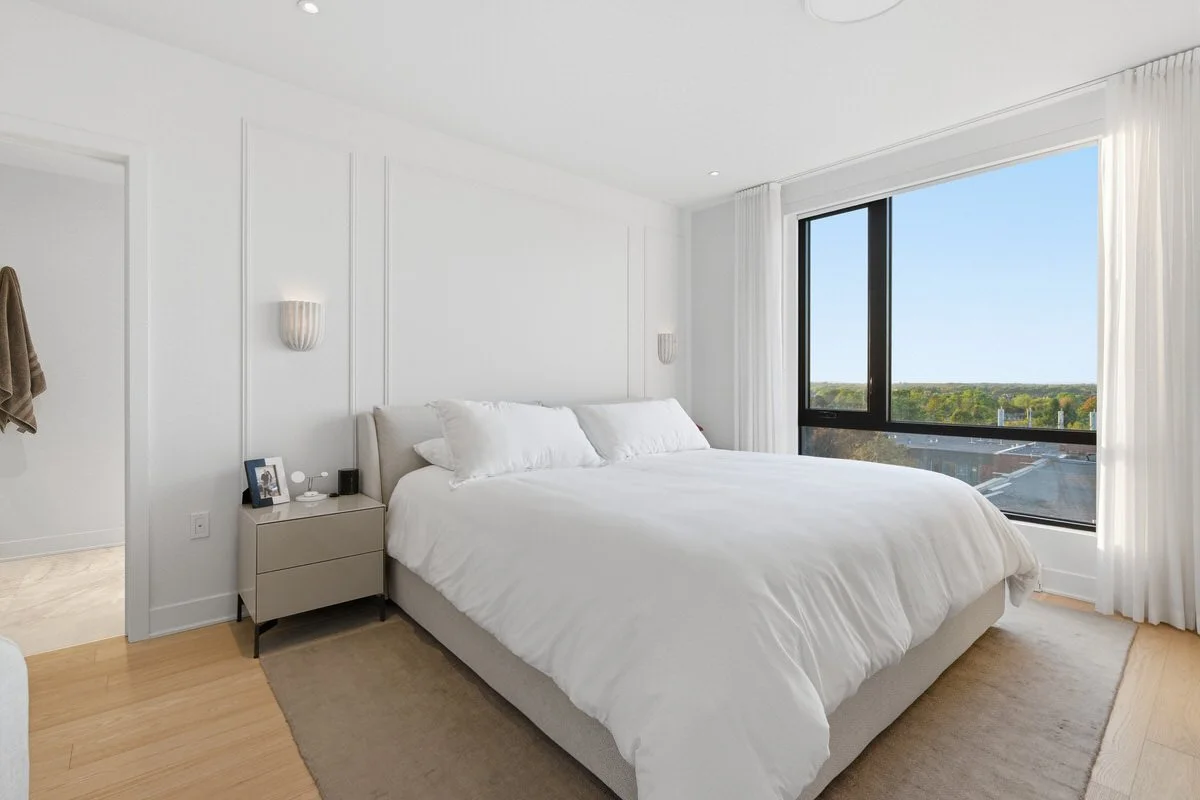 Bright bedroom with white bedding, nightstand with photo frame, clock, and lamp, large window with white curtains revealing a city view.