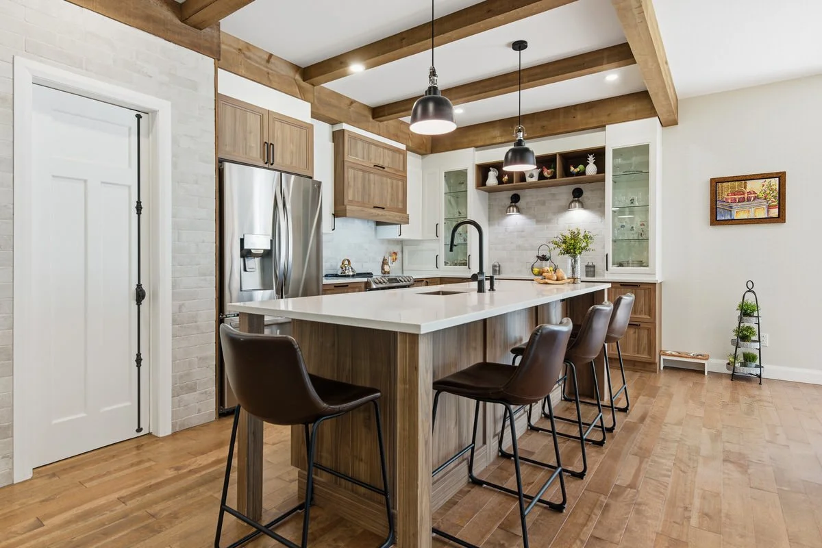 Modern kitchen with wooden cabinets, stainless steel refrigerator, white island with bar stools, glass-fronted cabinets, and decorative items on open shelves.
