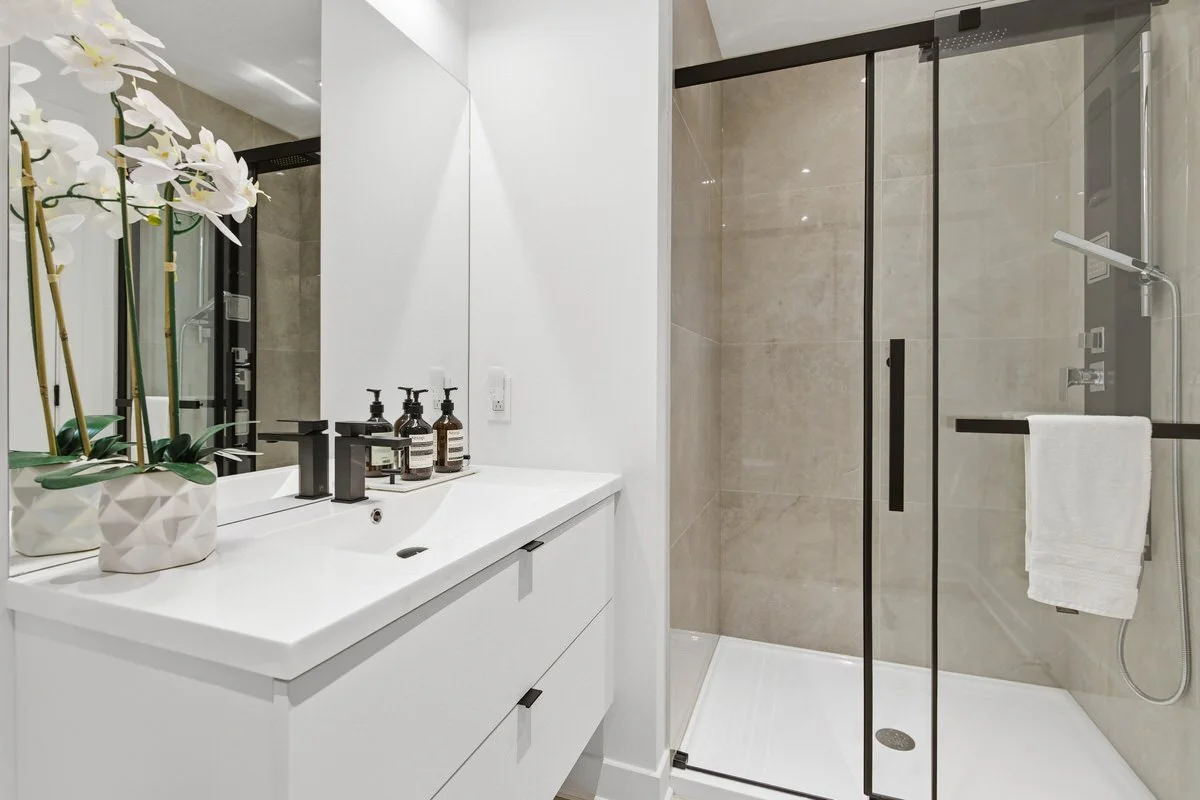 Modern bathroom with white vanity, potted orchid plants, soap dispensers, and a clear glass shower enclosure with black hardware and beige tile walls.