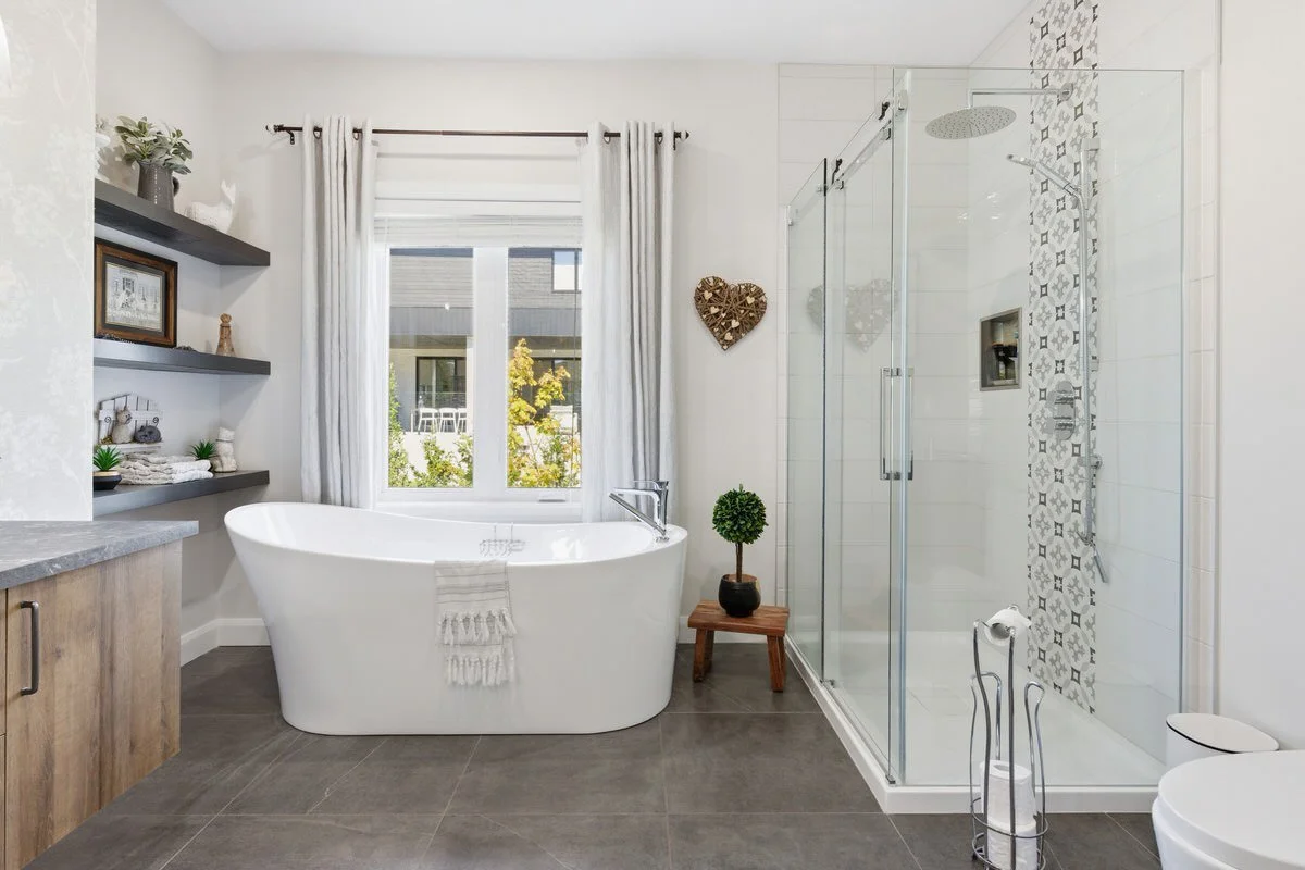 Modern bathroom with a white soaking tub, glass shower enclosure, decorative wall tiles in the shower, window with white curtains, wall shelves with decorative items, potted plant, and a wooden stool.