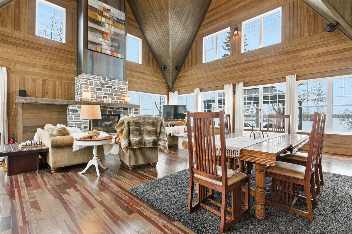 Open-concept living and dining room with wood-paneled walls and ceiling, large windows with white curtains, a stone fireplace, a television, a beige sofa with a blanket, a small side table with a lamp, a dark wooden side table, a chessboard, and a re