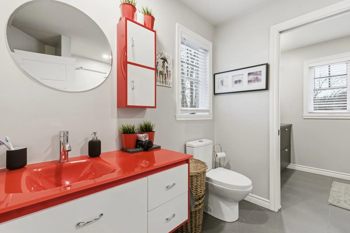 Modern bathroom featuring a red countertop sink, a round mirror, a vented cabinet above the sink, potted plants, framed art, a window with blinds, a toilet with a wicker basket, and a doorway to another room with a gray cabinet.