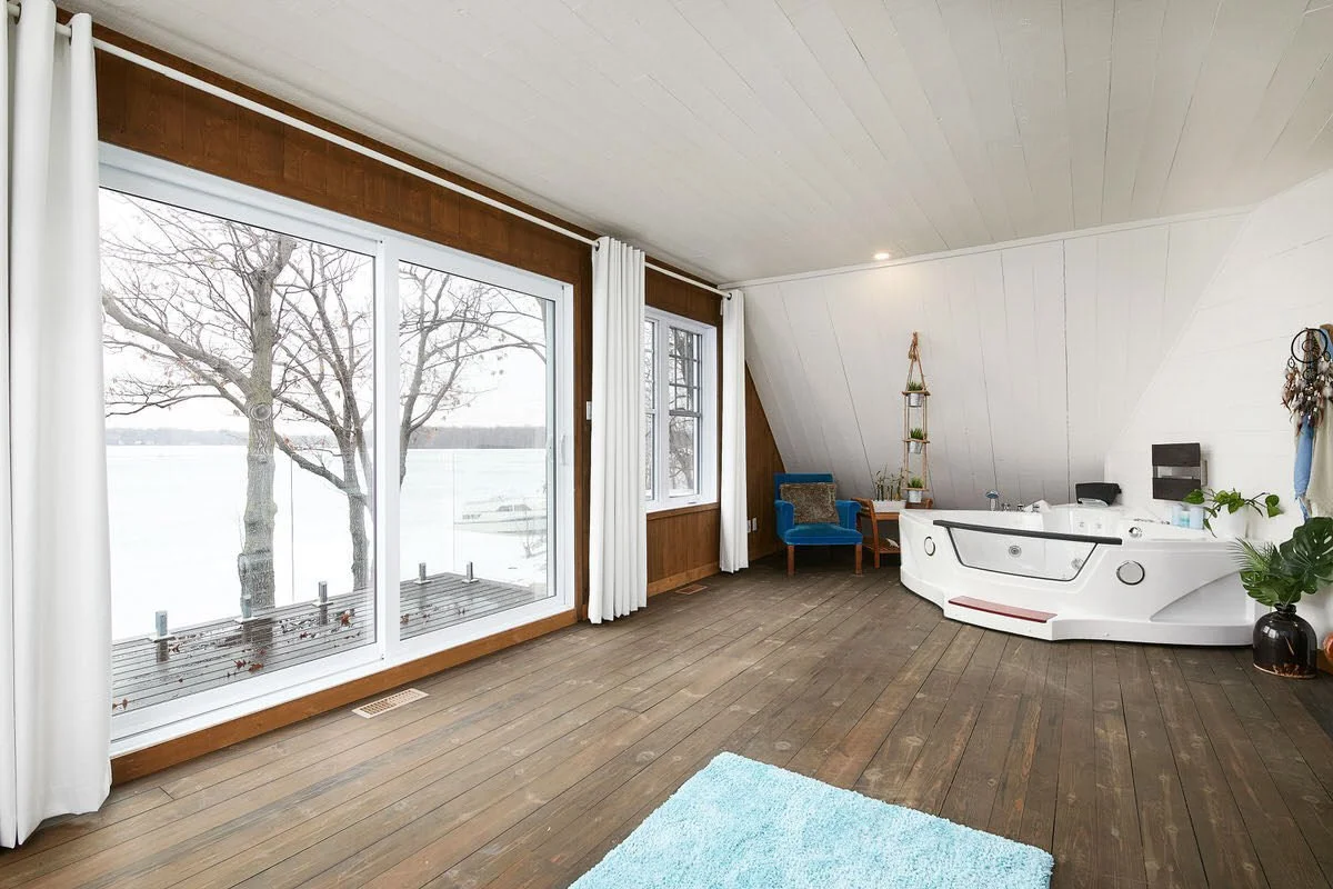 A spacious room with large windows overlooking a snowy outdoor scene with trees. The room features wooden floors, white curtains, a blue armchair, a small corner shelf with plants, and a white jacuzzi hot tub.