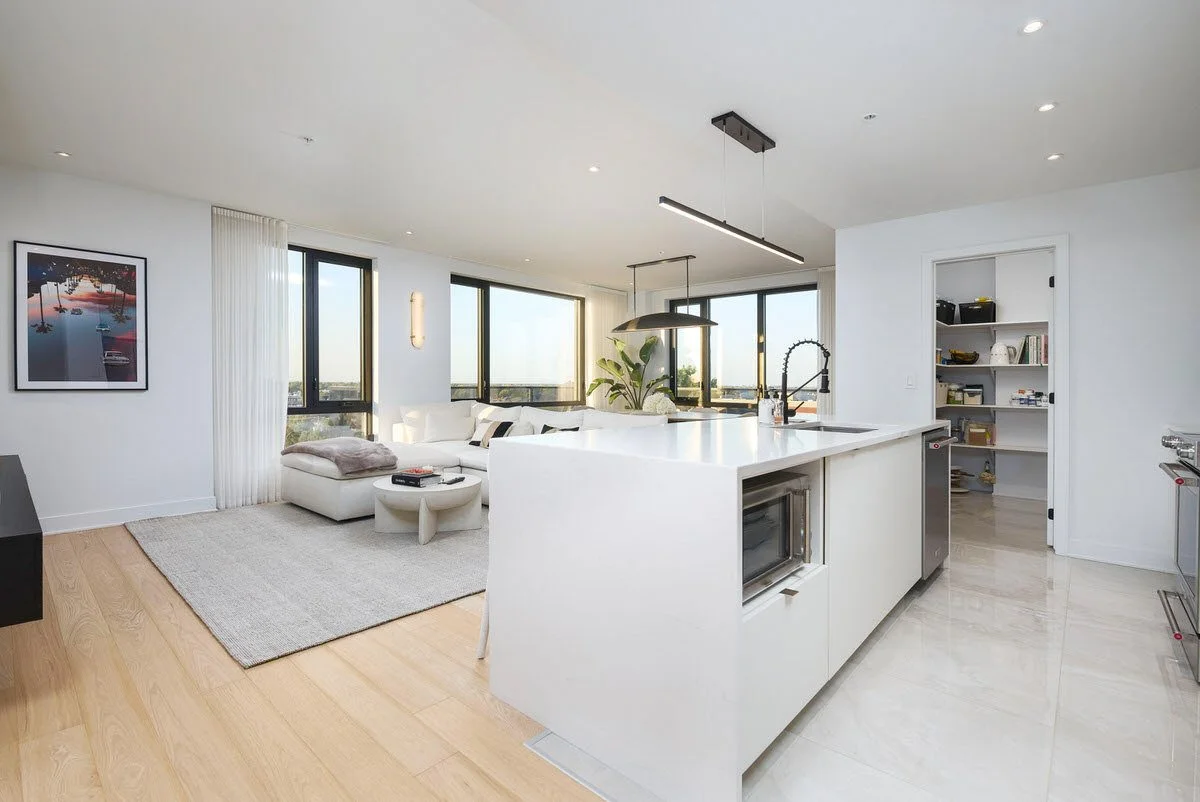 Modern open-concept living room and kitchen with large windows, white walls, minimalistic furniture, and natural light.