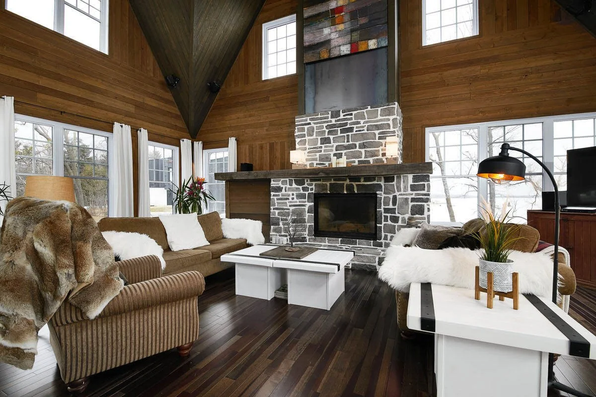 Living room with wooden walls, a stone fireplace, large windows with white curtains, a brown sofa with white pillows, a patterned armchair with a fur throw, a white coffee table, a side table with a plant, and a TV. A floor lamp with a black shade an