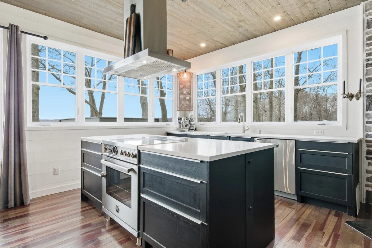 Modern kitchen with dark cabinets, white countertops, large windows, and a view of leafless trees outside, featuring a kitchen island and stainless steel appliances.