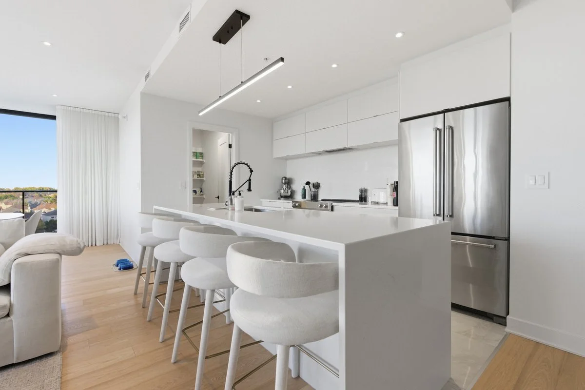 Modern white kitchen with a large island and four white bar stools, stainless steel refrigerator, and minimal decor, opening to a living area with a beige sofa and large window with white curtains.