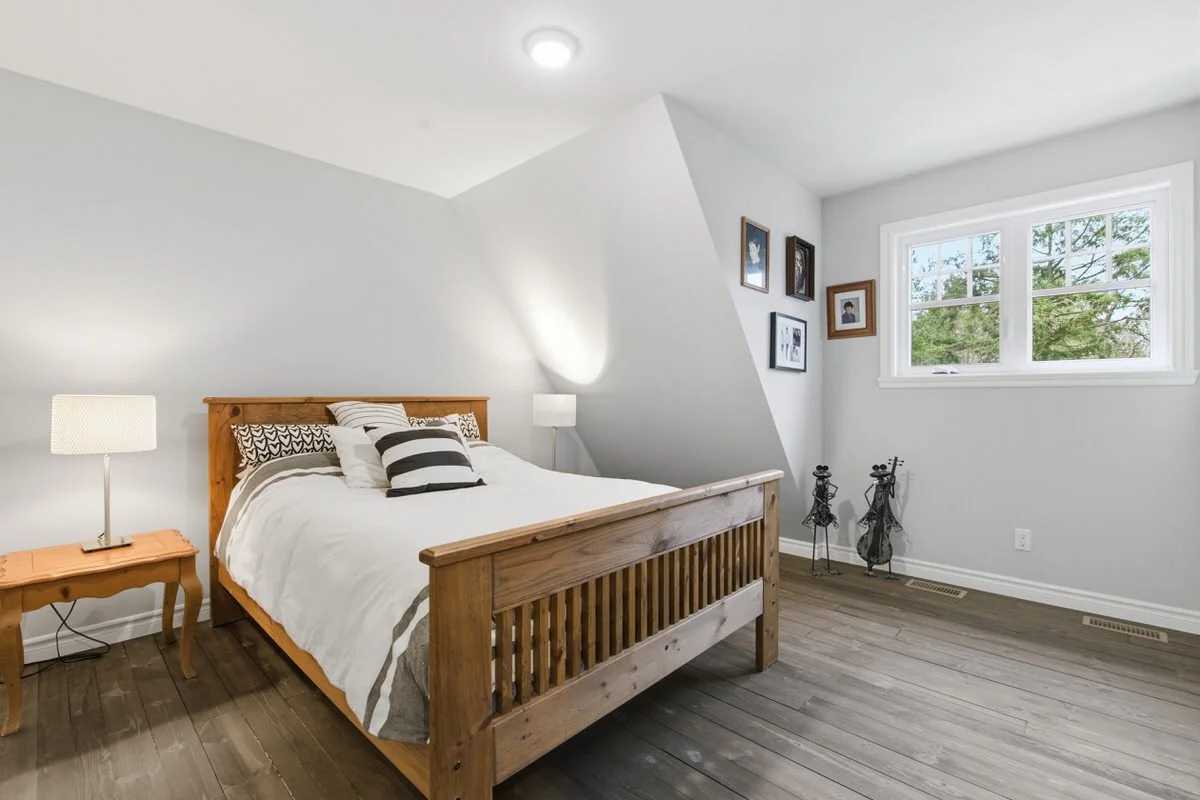 A bedroom with a wooden bed, white bedding, a nightstand with a lamp, framed pictures on the wall, and a window showing greenery outside.