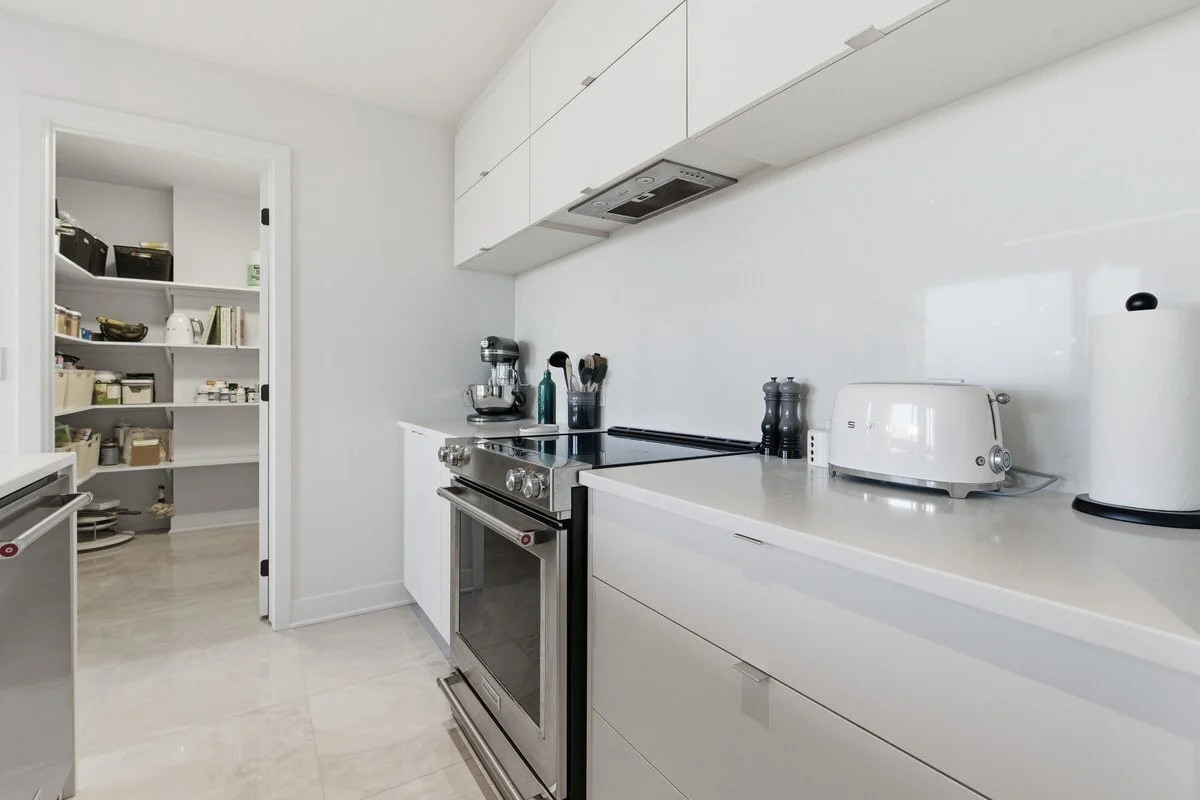 A modern, minimalistic white kitchen with a stainless steel oven, a stand mixer, a toaster, and a pantry with shelves filled with food and kitchen supplies.