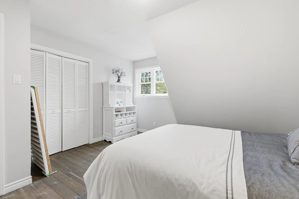 A minimalist bedroom with a sloped ceiling, white walls, and wooden floor. Contains a bed with a white and gray duvet, a white dresser with a small plant on top, and a white closet with louvered doors. A window with a view of trees is near the dresse