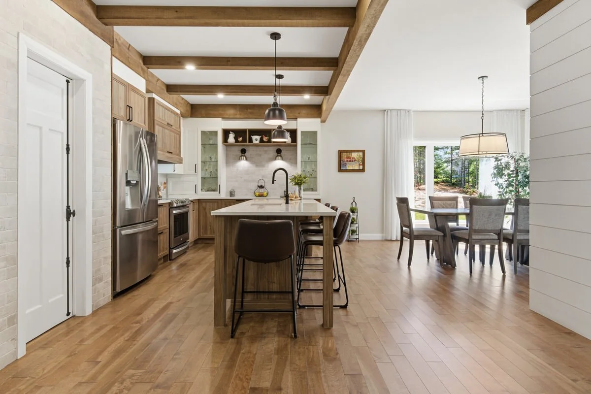 Open-concept kitchen with wooden cabinets, an island with barstools, stainless steel appliances, and a dining area with a table and chairs near large windows with white curtains.