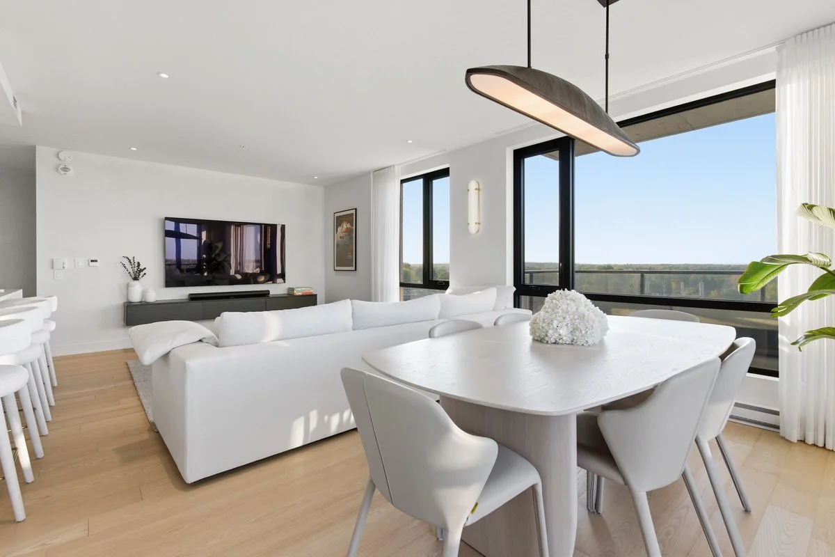 Bright living room with white sofa, dining table with white chairs, large windows with views, and modern lighting fixtures.