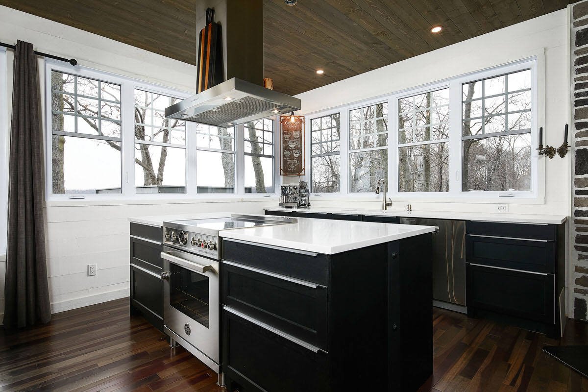 Modern kitchen with large windows, black cabinets, and a white countertop island, with a view of trees outside.